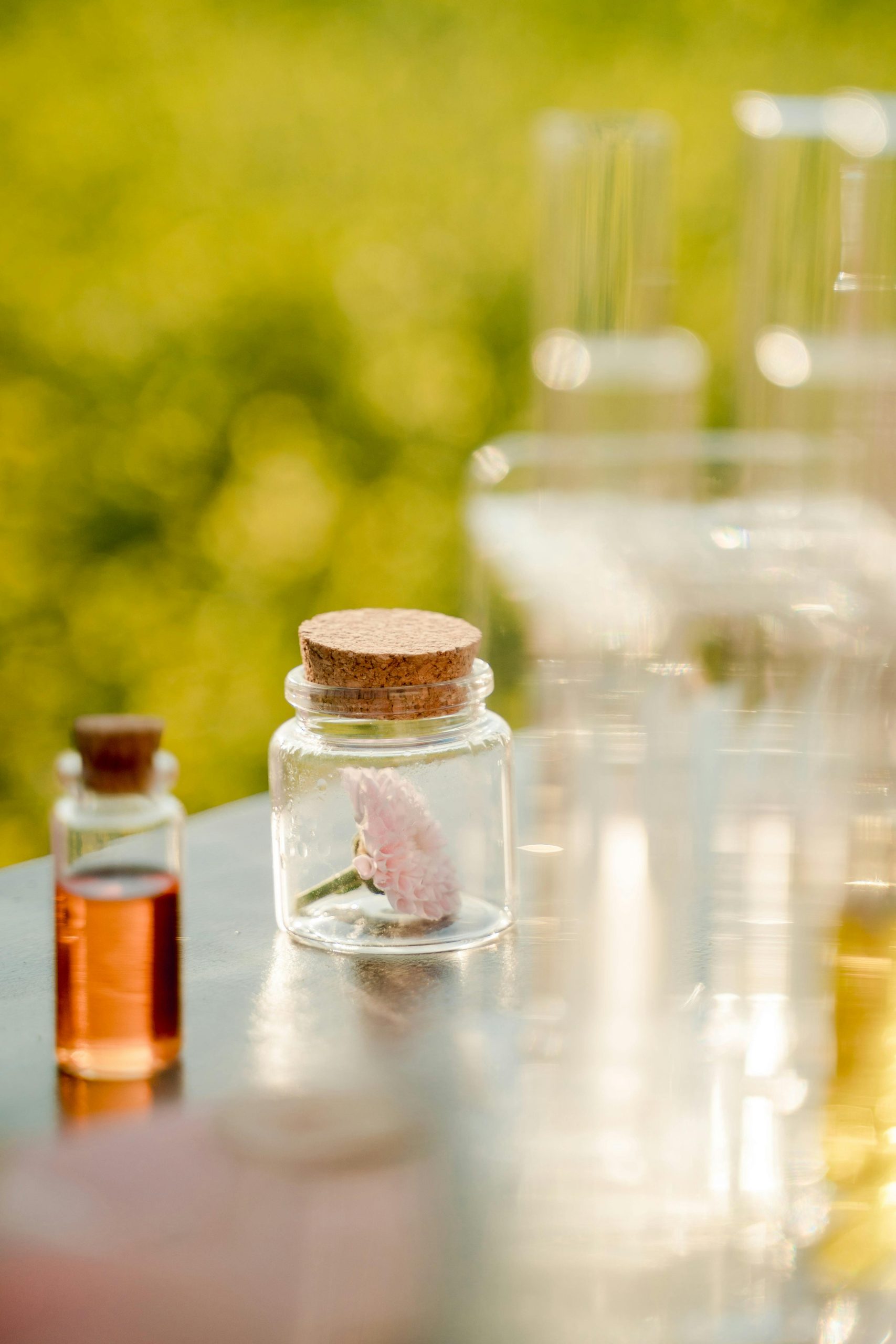 A close-up of glass bottles on a reflective surface with a flower in focus against an outdoor backdrop.